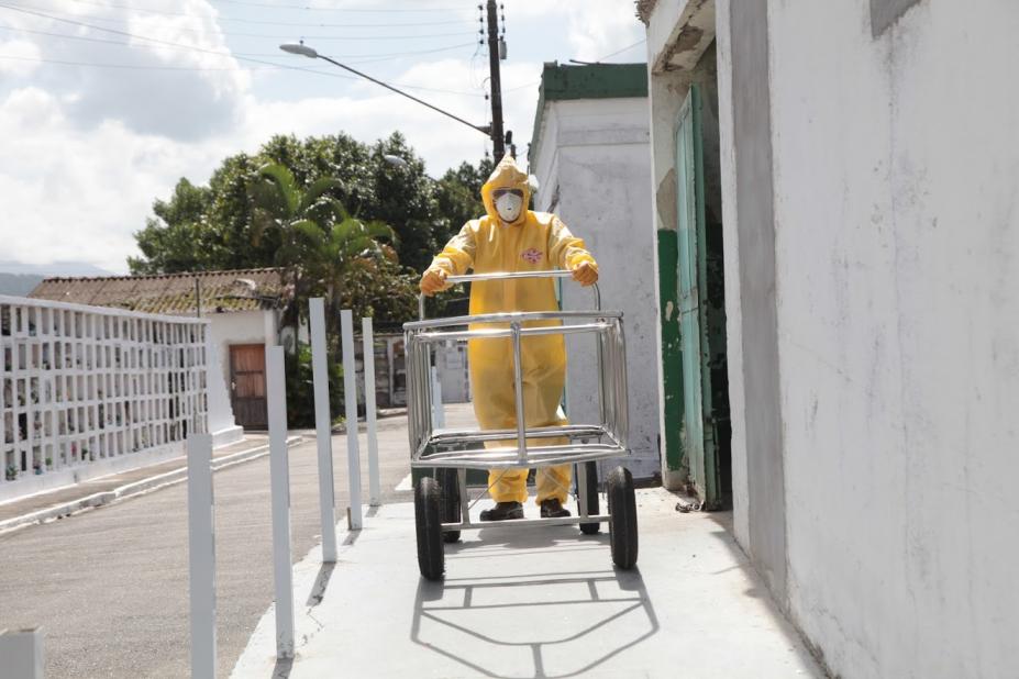 Funcionário vestindo macacão, luvas, máscara, gorro e botas empurra carrinho funerário vazio. #Paratodosverem