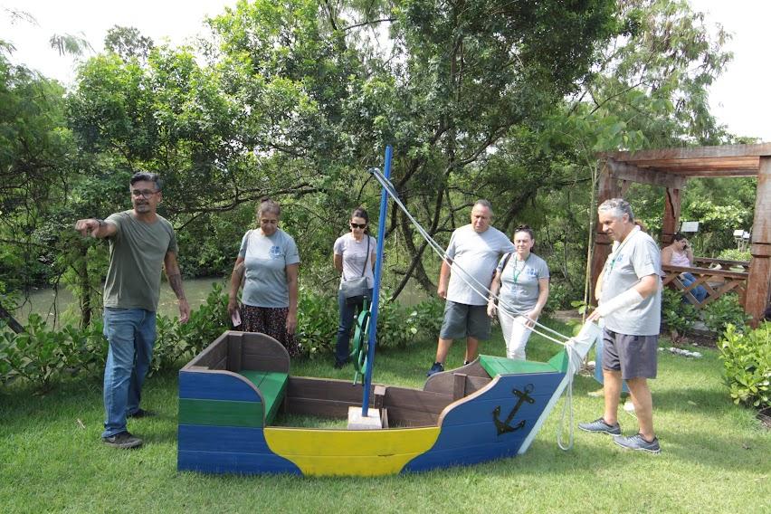 voluntários está em pé em um gramando olhando para uma peça em madeira, uma miniatura de barco. #paratodosverem