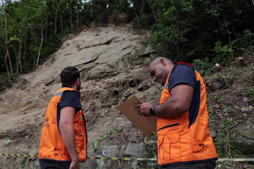 dois agentes da defesa civil estão na parte de baixo de um morro e à frente de uma encosta. Um deles faz anotações em uma prancheta. #paratodosverem