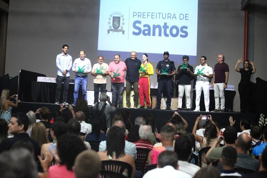 salão lotado de pessoas com palco ao fundo onde estão diversos atletas com seus troféus. #paratodosverem 