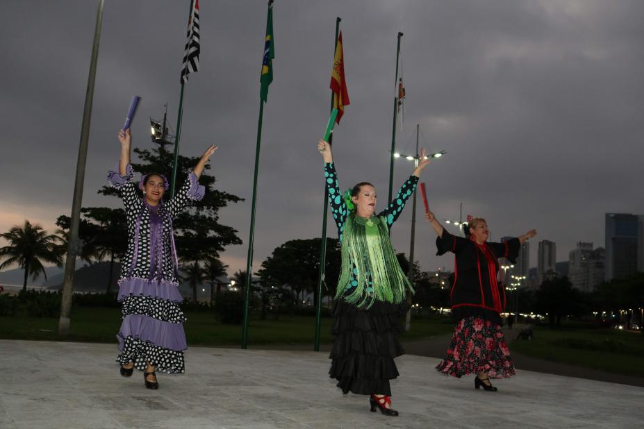 Três mulhes vestidas como dançarinas espanholas dançam à frente das bandeiras de Brasil, Espanha, São Paulo e Santos