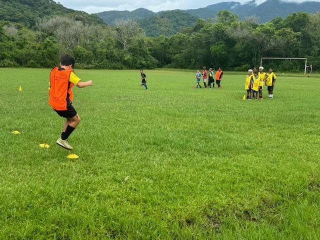 criança dá saltos em gramado em treino de futebol. Ao fundo há outras crianças e uma trave de gol mais ao fundo. #paratodosverem 