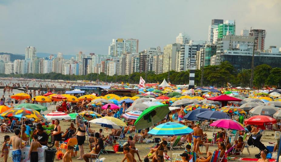 praia lotada em dia de sol com muitos guarda-sóis armados. #paratodosverem 