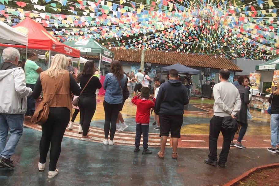 várias pessoas numa braça com barracas de comida e docorada com bandeirinhas no alto. #paratodosverem