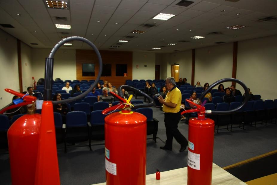 Instrutor conversa com público em auditório com três extintores de incêndio em primeiro plano. #pratodosverem