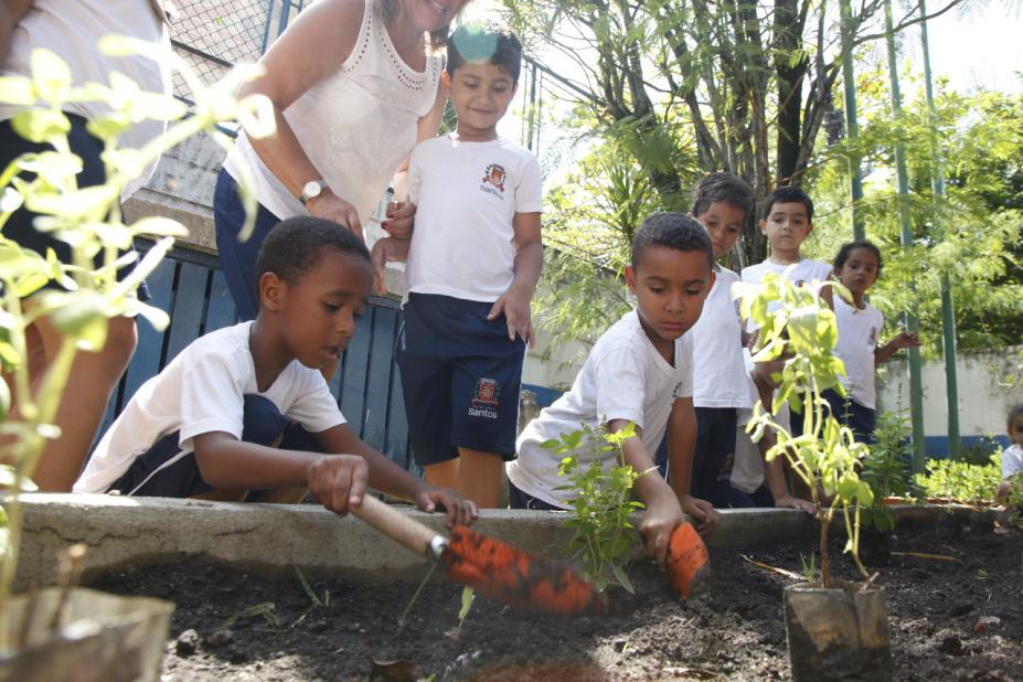 estudantes uniformizados estão mexendo em canteiro de plantas com pequenas pás. #paratodosverem