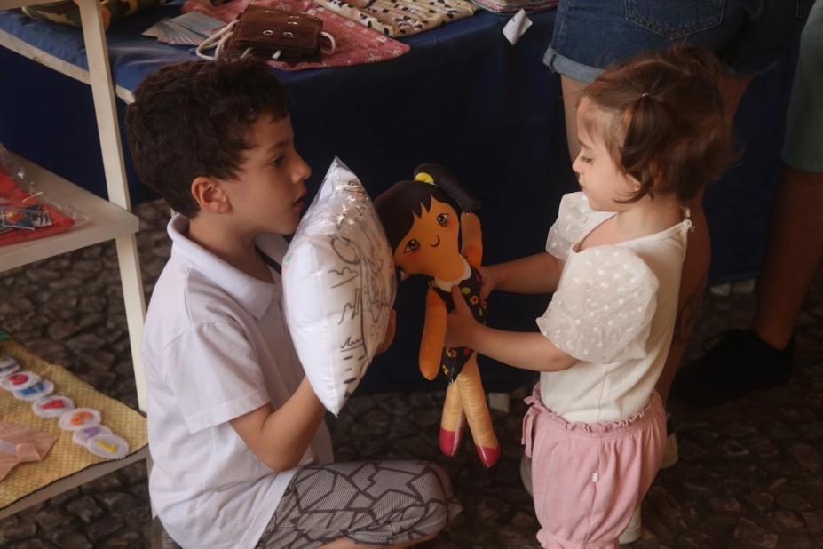 duas crianças estão frente a frente. O menino segura uma almofada e a menina, uma boneca de pano. #paratodosverem
