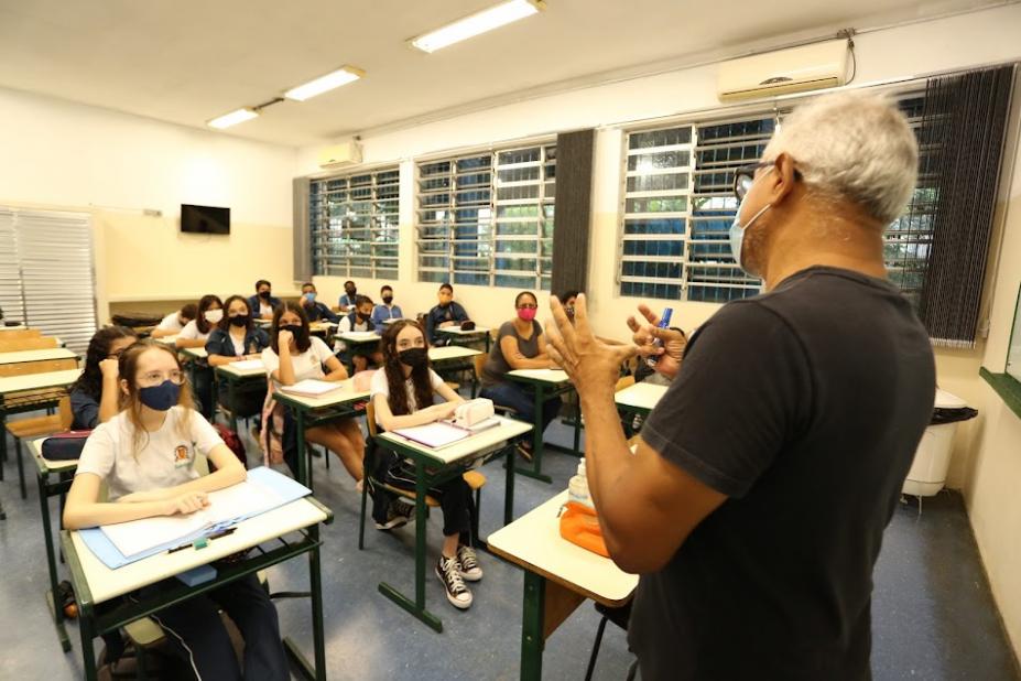 professor está à frente dos alunos e de costas para a foto. Alunos estão sentados, todos usando máscara e distanciados. #paratodosverem