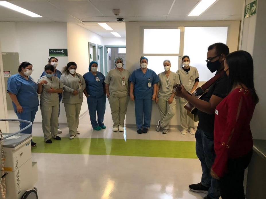 #pracegover Homem com violão e mulher se apresentam perante nove profissionais de saúde em corredor hospitalar