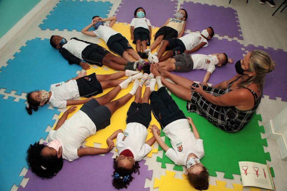 Crianças deitadas em círculo, juntas com professora, com todos encostando os pés. #pratodosverem