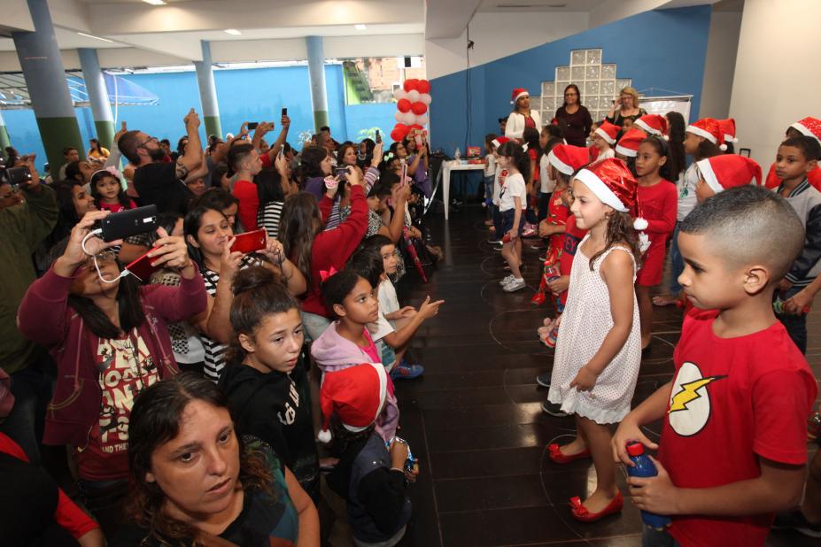 Crianças e adultos assistem apresentação de outras crianças que estão usando capuz de papai noel. #Pracegover