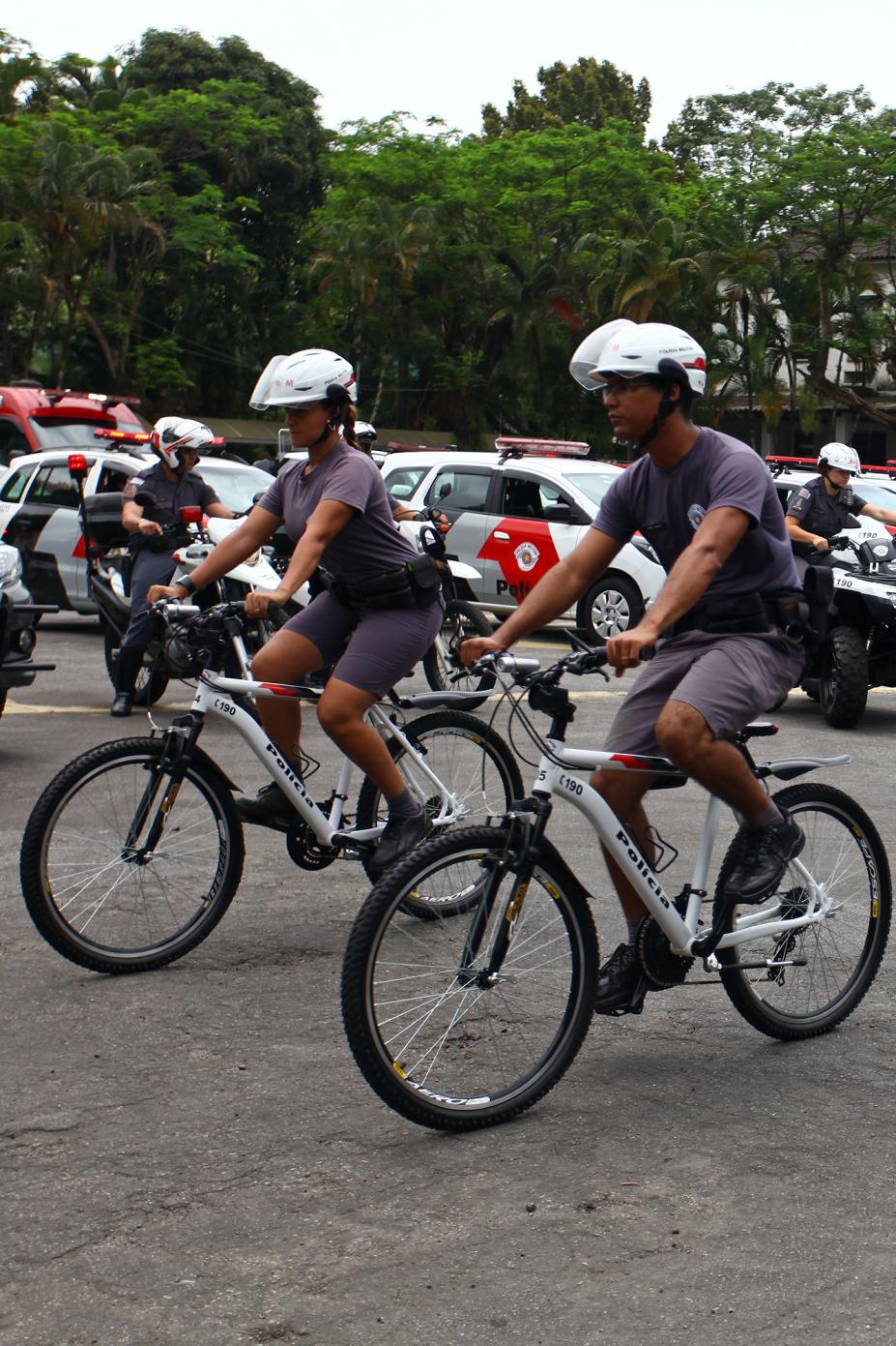 Dois policiais estão andando de bicicleta. Ao fundo há viaturas e policiais em motocicletas. #Pracegover