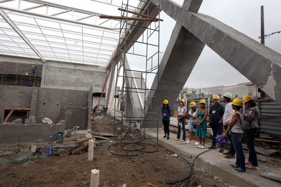 Pessoas estão em pé e paradas em uma obra. Elas estão abaixo de uma grande estrutura de concreto