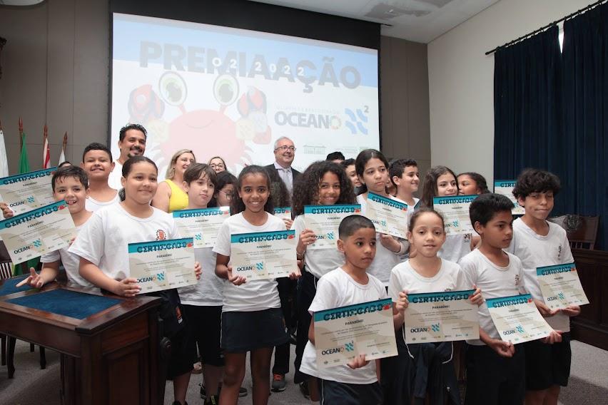 crianças posam para foto segurando certificados. Ao fundo há um telão onde se lê premiação. #paratodosverem