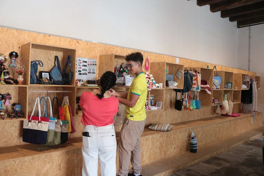 homem e mulher estão de costas para a foto e de frente para prateleira onde estão expostos produtos artesanais. #paratodosverem 