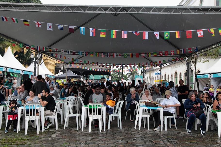 pessoas estão sentadas em praça de alimentação decorada com bandeirinhas de diversos países. #paratodosverem 