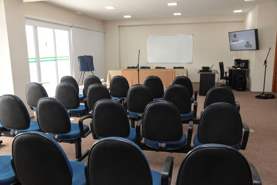 sala com diversas cadeiras e telão fixado ao fundo. #paratodosverem