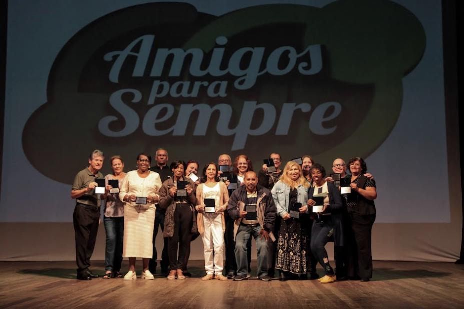 servidores reunidos no palco segurando medalhas posam para foto. Ao fundo, na parede se lê amigos para sempre. #paratodosverem