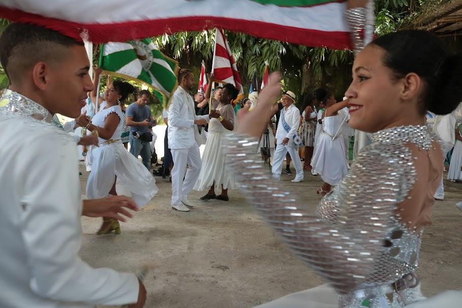 close de casal de mestre-sala e porta-bandeira com outros casais dançando ao fundo. #paratodosverem