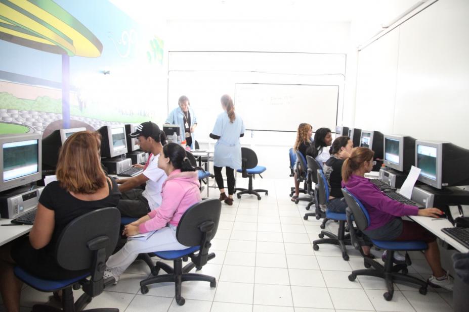 Sala de aula com diversos alunos diante de bancadas com computadores. #Pracegover