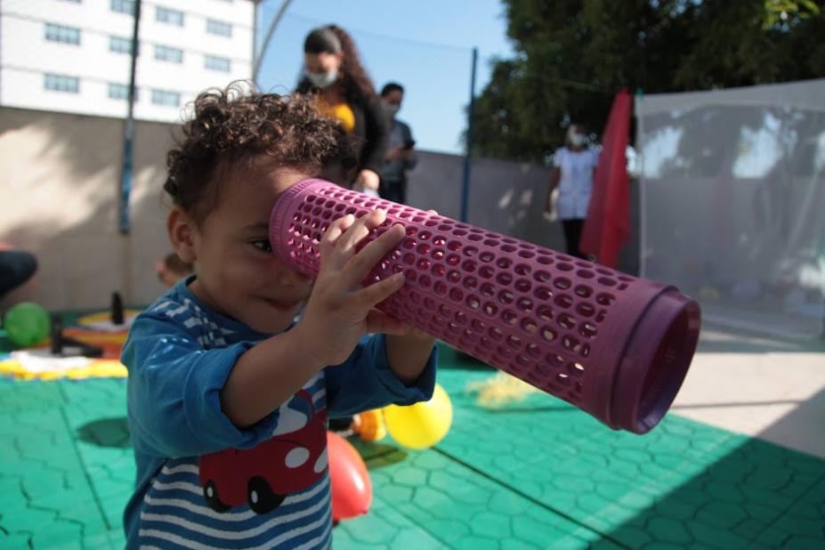 criança olha por dentro de tubo colorido cheio de furos. #paratodosverem