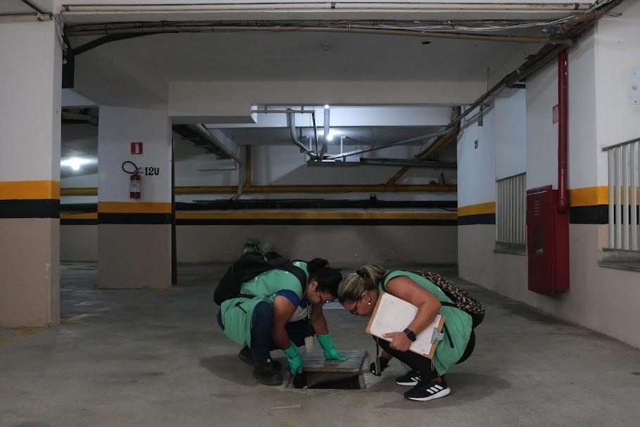 duas agentes uniformizadas agachadas examinam um ralo numa garagem de prédio. #paratodosverem 