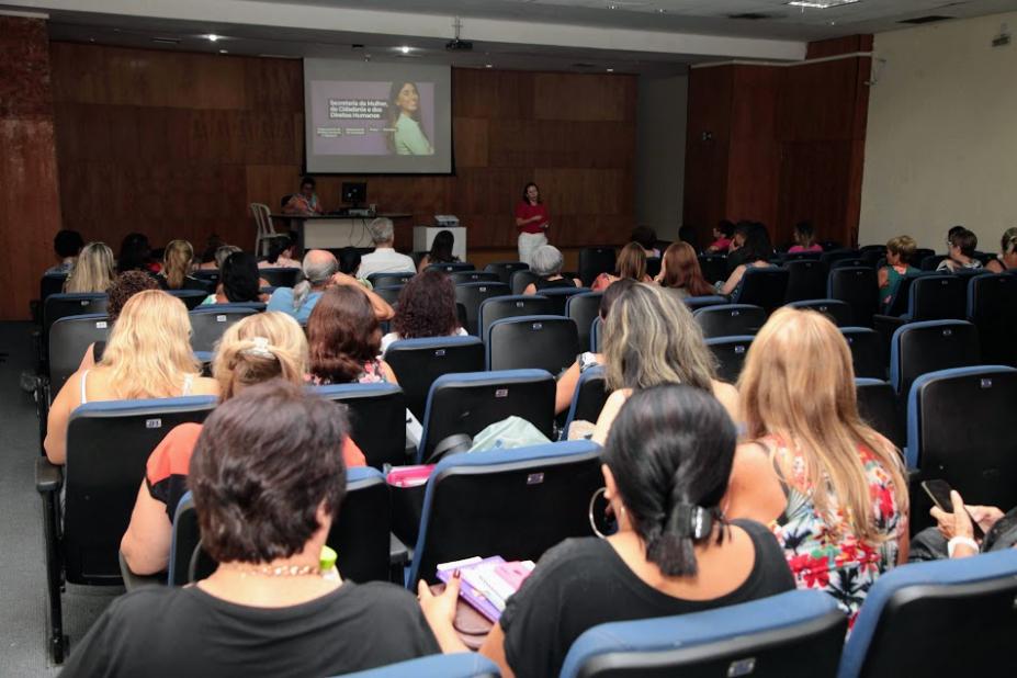 várias pessoas sentadas em um auditório e uma pessoa falando ao fundo à frente de um telão. #paratodosverem