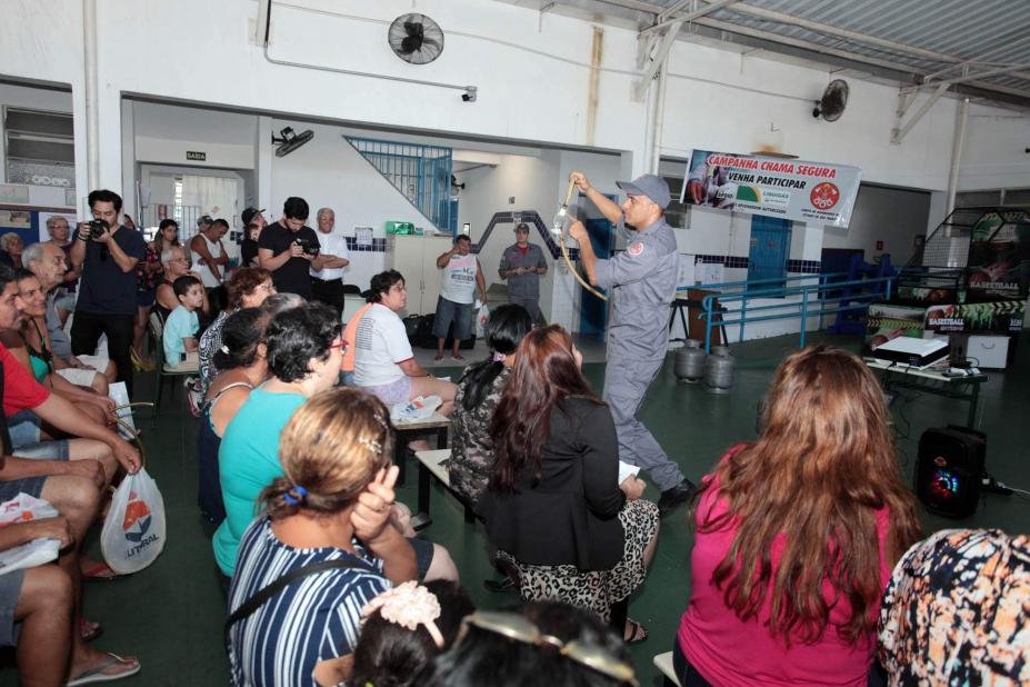 Cerca de 20 pessoas sentadas observam bombeiro uniformizado que mostra mangueira e válvula de botijão de gás