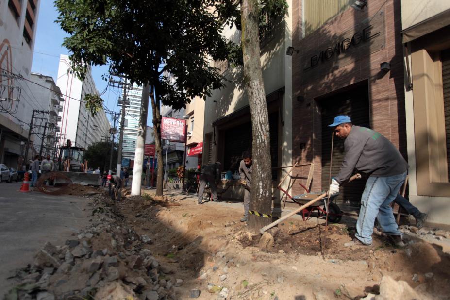 Homem trabalha na reforma de calçada de rua. Há pedras soltas e terra aparente. #Pracegover