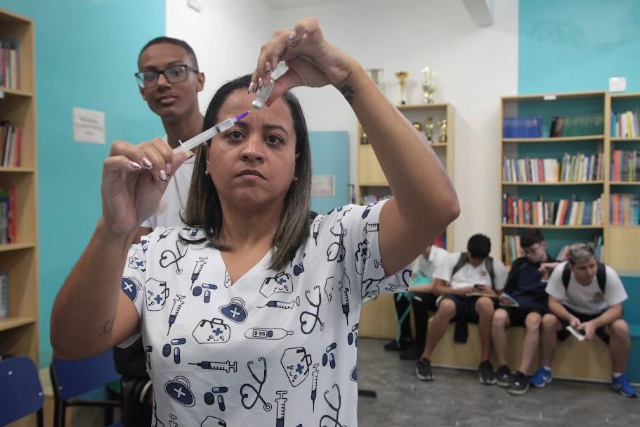 mulher prepara vacina em seringa. Há um jovem em pé atrás dela. e outros sentados mais ao fundo. É uma sala escolar. #paratodosverem