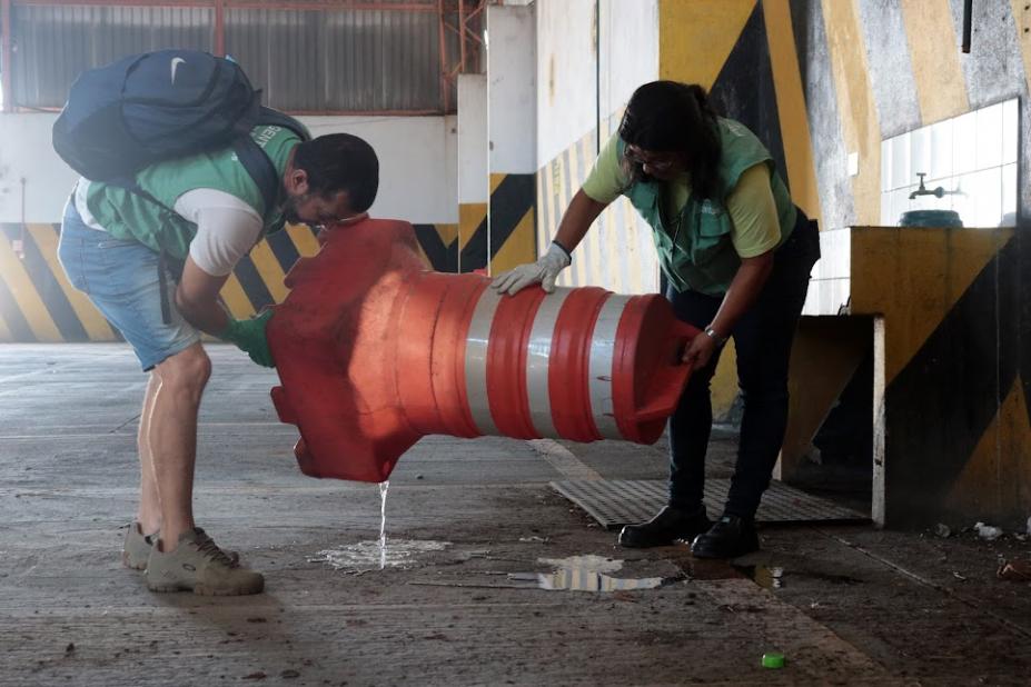 Dois agente seguram cone de trânsito para despejar água contida no interior. #paratodosverem 