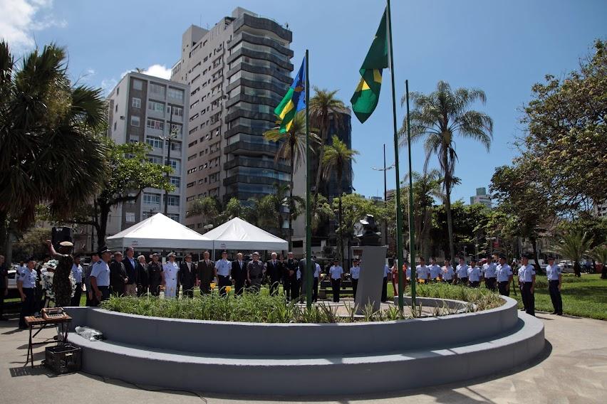 duas bandeiras hasteadas em canteiro no jardim da orla. Várias pessoas estão em pé sob tendas em cerimônia. #paratodosverem