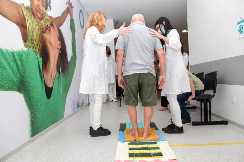 idoso caminha entre duas mulheres que usam avental branco. #paratodosverem