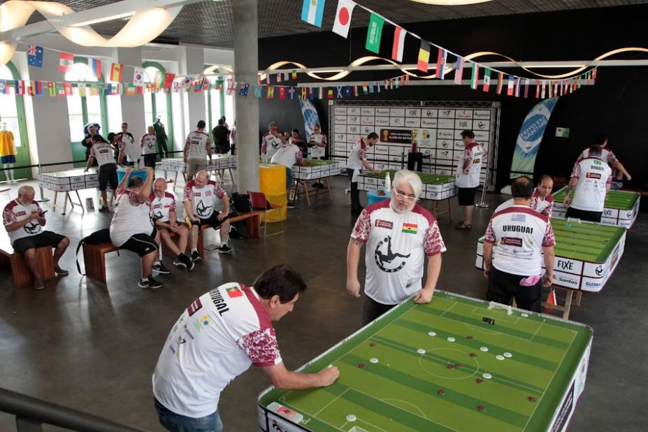 várias duplas jogando futebol de botão no Museu Pelé decorado por bandeiras de times participantes da copa. #paratodosverem