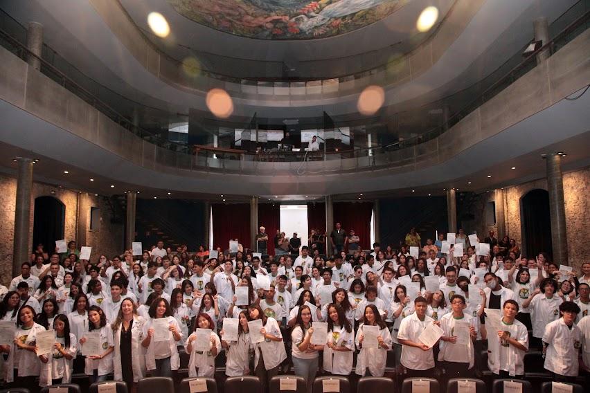 VÁRIOS JOVENS USANDO UNIFORME E AVENTAL  BRANCO NA PLATEIA DE TEATRO. tODOS SEGURAM SEUS CERTIFICADOS