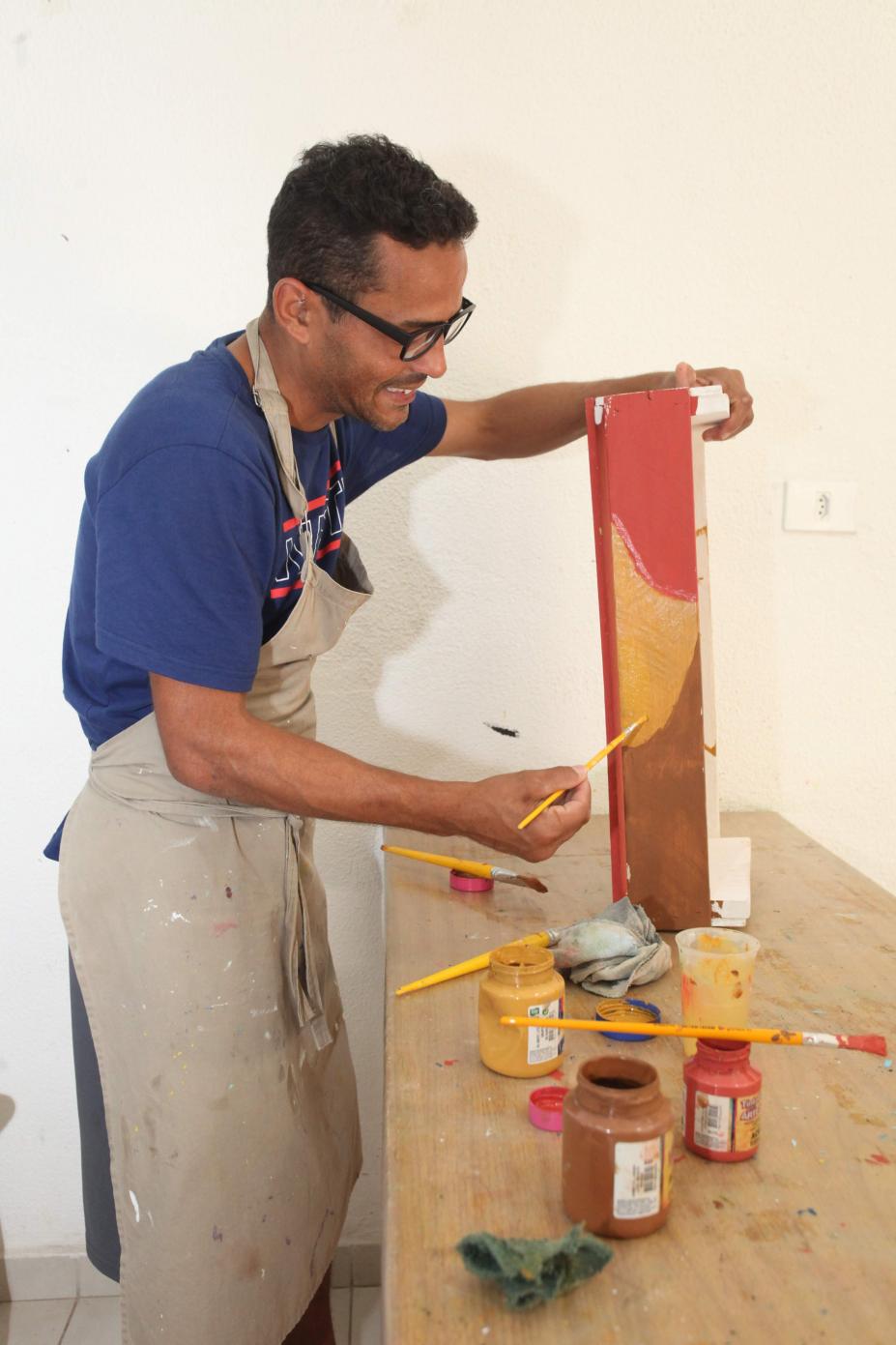 Homem está pintando peça de madeira. Há potes de tinta e pinceis sobre a mesa. #Pracegover