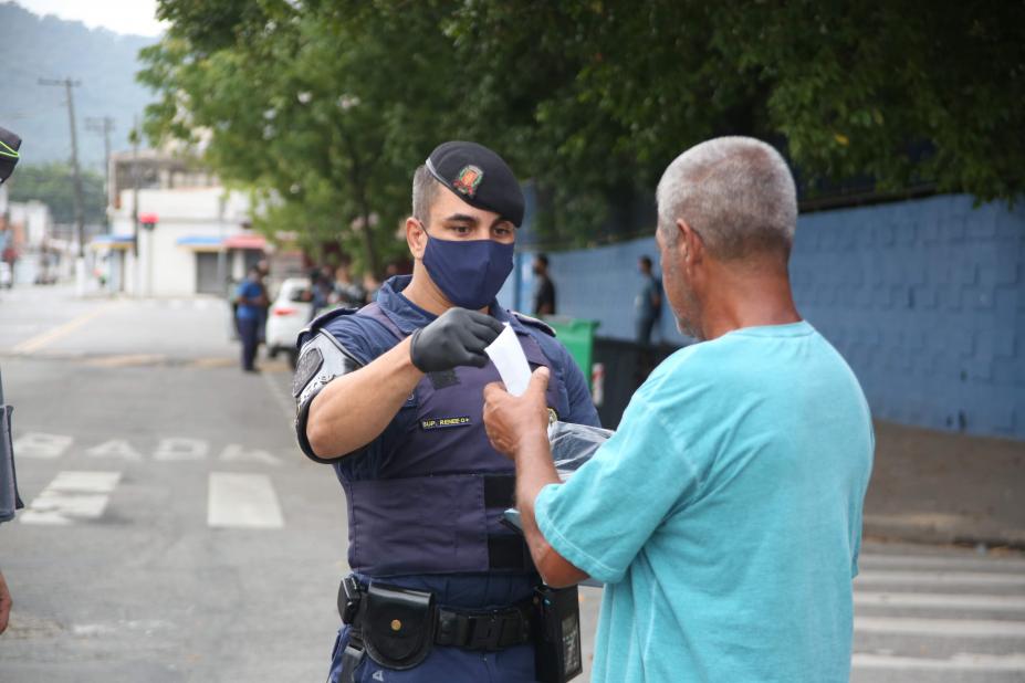 #pracegover Com máscara no rosto, guarda municipal entrega máscara descartável a idoso em calçada de avenida