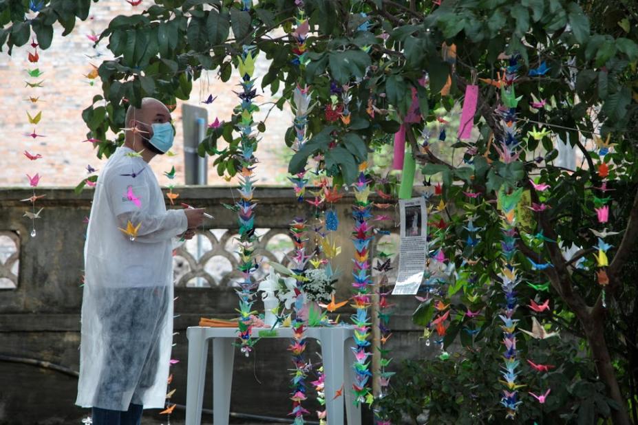 Enfermeiro com caneta e papel na mão observa árvore cheia de origamis coloridos. #paratodosverem