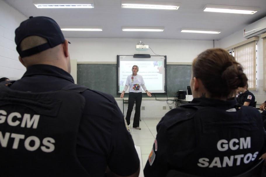 dois guardas municipais com coletes, sentados, de costas para a foto. Ao fundo, aparecendo entre o meio dos ombros deles dois, há uma mulher falando à frente de um telão. #paratodosverm