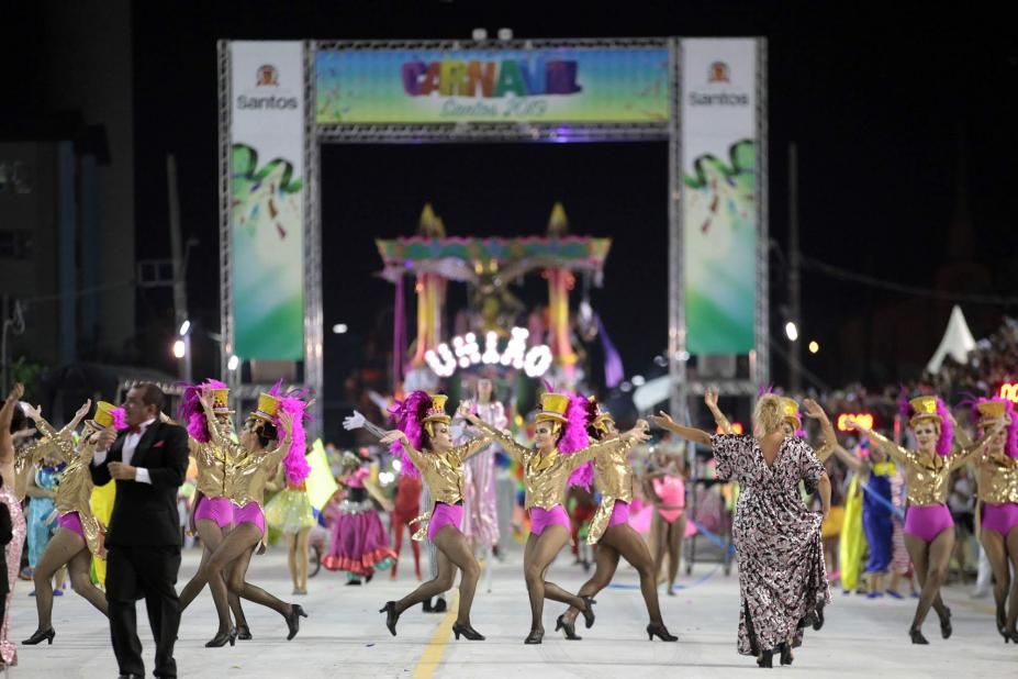 Escola de samba está desfilando na avenida. Ao fundo, o pórtico de entrada, onde se lê Carnaval. #Pracegover