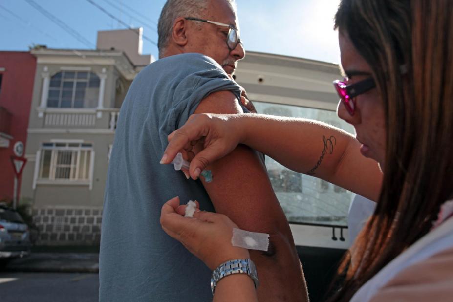 Agente da saúde vacina homem. Ao fundo se vê alguns sobrados na rua. #Pracegover