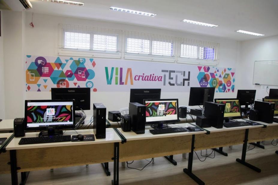 mesas com desktops em cima. Ao fundo, na parede, uma faixa onde se lê Vila Criativa Tech. #paratodosverem