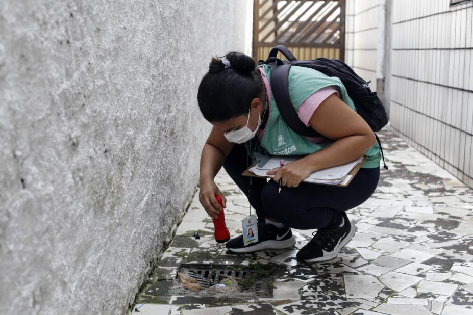 mulher vestindo colete verde agachada olhando um ralo externo #paratodosverem