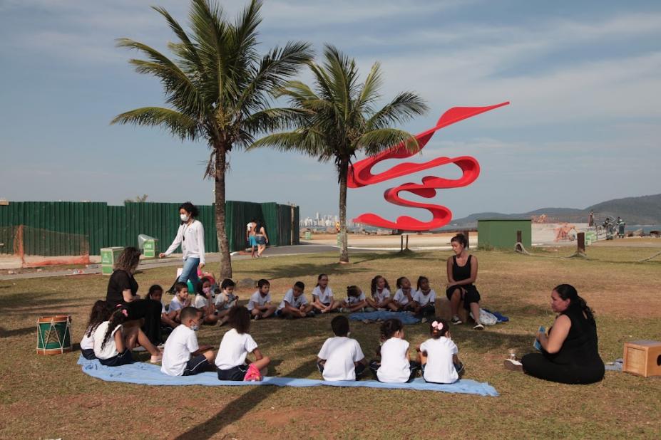 criancas sentadas na grama com escultura ao fundo em paruqe #paratodosverem
