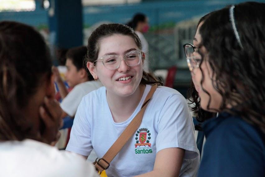 Bruna está sentada, de frente para a foto, conversando com amigas que aparecem de costas para a foto. #paratodosverem