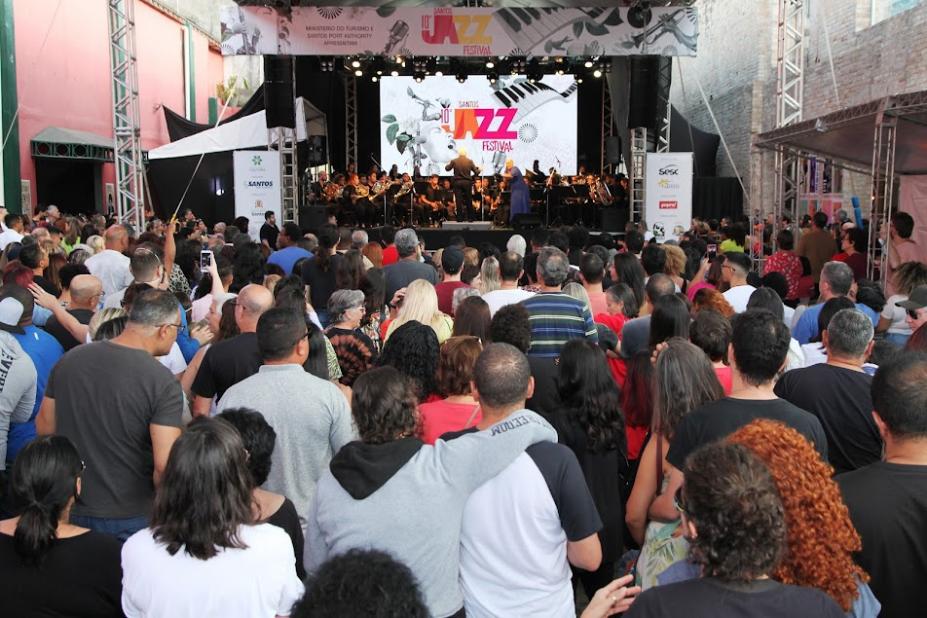 grande público assiste apresentação musical no palco ao fundo. #paratodosverem