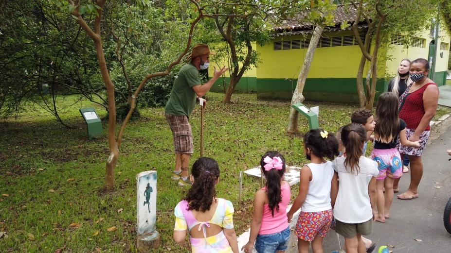 crianças estão no jardim botânico, de frente para um homemm que está à frente de uma árvore.Ele fala com todas elas. #paratodosverem