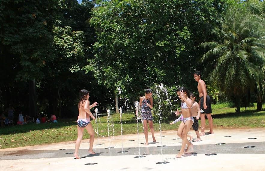 fonte interativa com crianças brincando. Vasta vegetação ao fundo. #paratodosverem