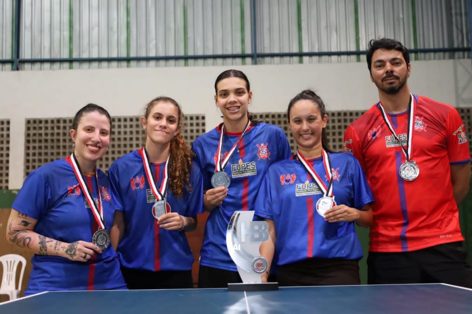 quatro atletas do tênis de mesa feminino seguram suas medalhas e posam atrás do troféu da Cidade. #paratodosverem