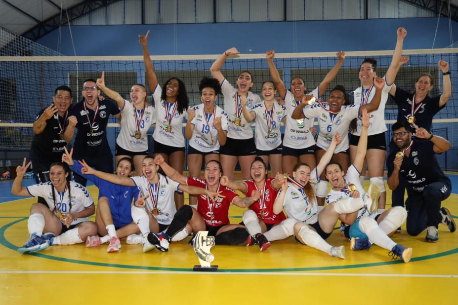 equipe feminina de vôlei reunida na quadra, com todas comemorando e posando para foto. #paratodosverem